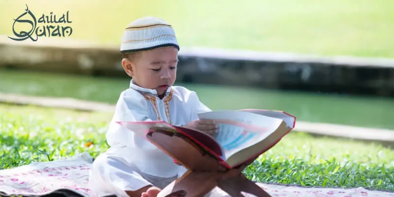 A child in a white thobe and kufi sitting on a patterned blanket.